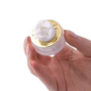 Hand holding a small container of white cream with a gold lid on a white background